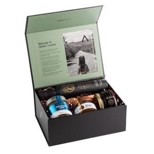 May include: A black gift box with a light green interior, containing olive oil, honey, and other gourmet food items. The box includes a photo and text that reads "Welcome to Geraki, Laconia."