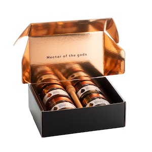 May include: A black gift box with a copper-colored interior holds several small jars of honey and a wooden honey dipper. The box's interior reads "Nectar of the gods". The jars are labeled "Raw Honey" and "Wildflower Honey".