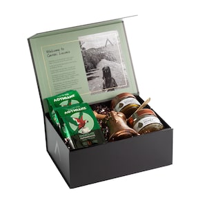 May include: A black gift box with a hinged lid, containing green packages of coffee, two jars of honey, and a bronze bell. The box's interior features a printed image and text. The coffee packages have a parrot illustration.