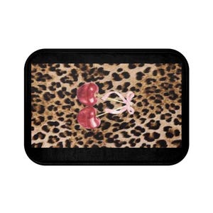 May include: A rectangular bath mat with a black border. The mat features a leopard print background with two red cherries tied together with a pink ribbon.