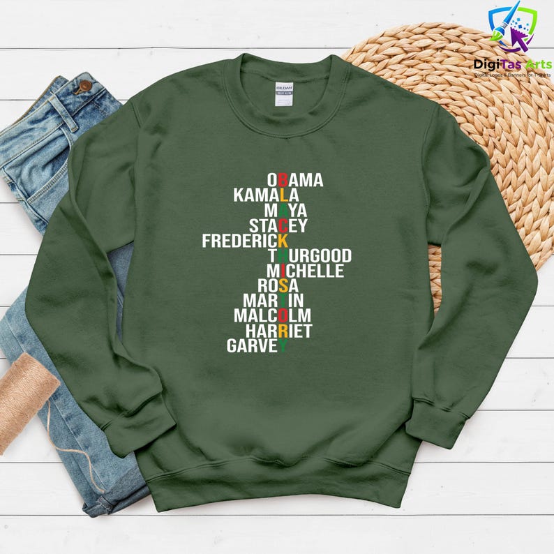 May include: A dark green sweatshirt featuring a list of names in white text, with the first letter of each name in Pan-African flag colours. The sweatshirt is displayed on a neutral surface. The names are stacked vertically.
