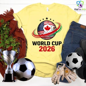Może przedstawiać: Ż&oacute;łta koszulka z motywem piłki nożnej z flagami Stan&oacute;w Zjednoczonych, Kanady i Meksyku, z napisem "WORLD CUP 2026". Obraz zawiera piłkę nożną, rękawice, trofeum i dżinsy.