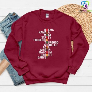 May include: A maroon sweatshirt featuring the names Obama, Kamala, Maya, Stacey, Frederick, Thurgood, Michelle, Rosa, Martin, Malcolm, Harriet, and Garvey in white, red, yellow, and green text.
