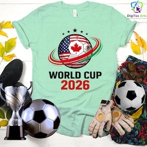 Może przedstawiać: Jasnozielona koszulka z motywem piłki nożnej z flagami USA, Kanady i Meksyku, z napisem "WORLD CUP 2026". Obraz zawiera piłki nożne, rękawice i trofeum.