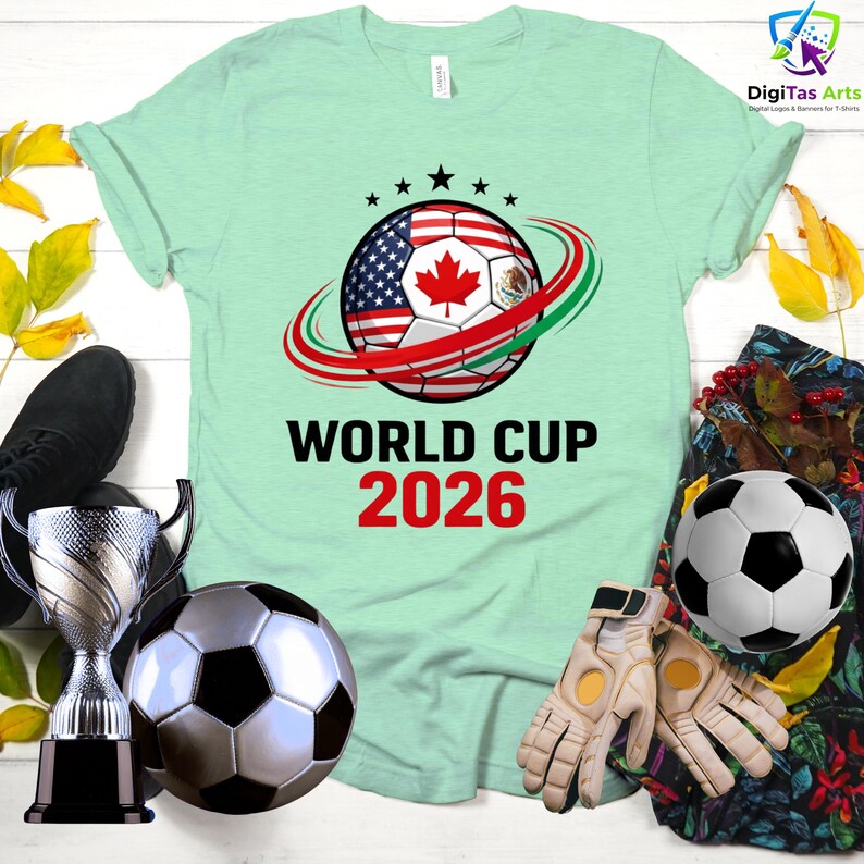Może przedstawiać: Jasnozielona koszulka z motywem piłki nożnej z flagami USA, Kanady i Meksyku, z napisem "WORLD CUP 2026". Obraz zawiera piłki nożne, rękawice i trofeum.
