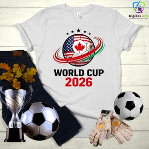 Może przedstawiać: Biała koszulka z nadrukiem piłki nożnej z flagami Stan&oacute;w Zjednoczonych, Kanady i Meksyku. Tekst "WORLD CUP 2026" jest wydrukowany poniżej w kolorze czarnym i czerwonym. Dodatkowo piłka nożna, trofeum i rękawice bramkarskie.