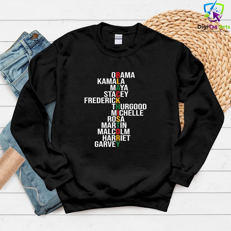 May include: Black crew neck sweatshirt featuring a list of names in white, red, yellow, and green text. The names include Obama, Kamala, Maya, Stacey, Frederick, Thurgood, Michelle, Rosa, Martin, Malcolm, Harriet, and Garvey.