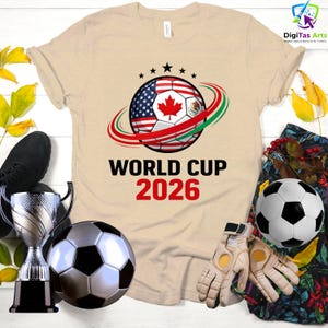 Może przedstawiać: Beżowa koszulka z motywem piłki nożnej z flagami USA, Kanady i Meksyku, z napisem "WORLD CUP 2026". Zawiera piłkę nożną, trofeum i rękawice.