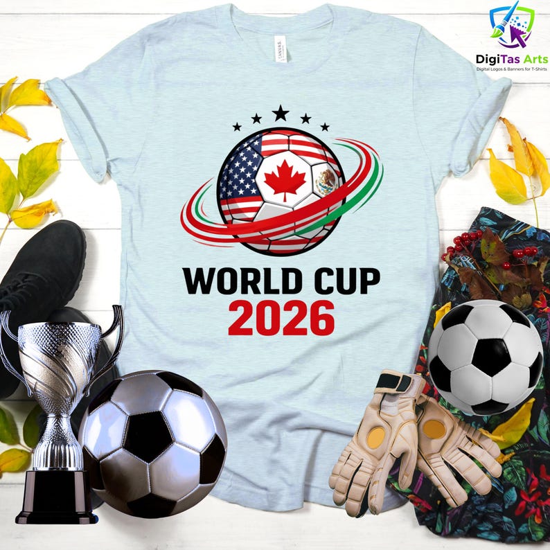 Może przedstawiać: Jasnoniebieska koszulka z motywem piłki nożnej, zawierającym flagi USA, Kanady i Meksyku, z napisem "WORLD CUP 2026". Na zdjęciu znajduje się r&oacute;wnież piłka nożna, trofeum, rękawice i czarny but.