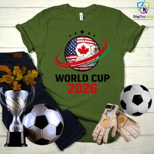 Może przedstawiać: Zielona koszulka z nadrukiem piłki nożnej z flagami USA, Kanady i Meksyku. Tekst "WORLD CUP 2026" jest wydrukowany w kolorze czarnym i czerwonym. Zawiera piłkę nożną, trofeum i rękawice.