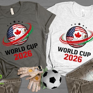 Może przedstawiać: Dwie koszulki, jedna szara i jedna biała, z motywem piłki nożnej z flagami USA, Kanady i Meksyku. Tekst "WORLD CUP 2026" jest wydrukowany poniżej w kolorze czarnym i czerwonym.