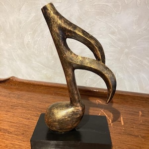 Könnte beinhalten: Eine Holzskulptur einer Musiknote (Achtelnote), bemalt in Gold- und Brauntönen. Die Note ist auf einem schwarzen rechteckigen Sockel montiert. Die Skulptur ist ein dekoratives Objekt.