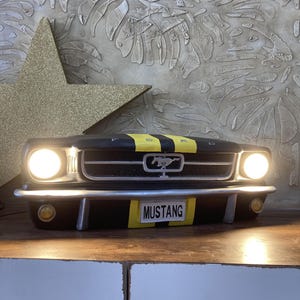 Puede incluir: Lámpara con forma de coche Mustang negro y amarillo con dos faros brillantes y una placa que dice "MUSTANG". La lámpara tiene una franja amarilla en el capó y está sobre una superficie de madera.