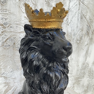 May include: A black lion statue wearing a gold crown. The lion has a detailed mane and is sitting upright. The crown is ornate with a textured surface. The background is a textured white and grey.