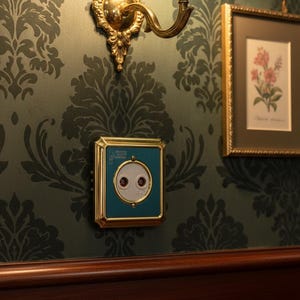 Könnte beinhalten: Eine türkisfarbene und goldfarbene Steckdose mit zwei roten Punkten, montiert an einer dunkelgrünen Wand mit Damasttapete. Eine goldene Wandleuchte und ein gerahmter botanischer Druck sind ebenfalls zu sehen.
