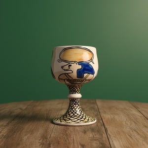 Puede incluir: Una copa de cerámica pintada a mano con base crema y tallo detallado. La copa presenta un diseño con detalles en azul y amarillo. El tallo tiene un patrón de escamas de pez. La copa está sobre una superficie de madera sobre un fondo verde.