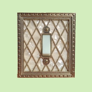 Puede incluir: Un interruptor de luz vintage con un marco decorativo de color dorado. El interruptor tiene una palanca blanca y un fondo con patrón de diamantes. El marco tiene un borde con cuentas y está montado con dos tornillos.