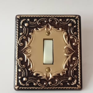 Puede incluir: Una placa de interruptor de luz rectangular con un borde marrón oscuro, adornos dorados en relieve y una placa central de metal dorado. Un interruptor de palanca blanco se encuentra en el centro, asegurado por dos pequeños tornillos dorados.