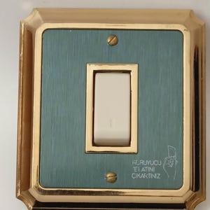 Puede incluir: Un interruptor de luz de estilo vintage con un marco dorado y un panel central de color turquesa. El interruptor es blanco y el panel tiene una textura de metal cepillado. El texto "KORUYUCU ELATINI CIKARTINIZ" es visible.