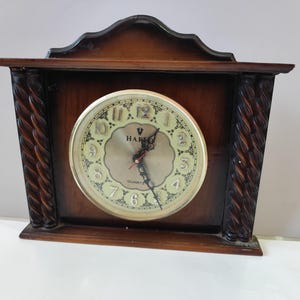 May include: A dark brown wooden mantel clock with a rectangular frame, a decorative arched top, and two vertical twisted columns on the sides. The circular clock face features a gold background, black Arabic numerals, black hands, and the text HARIO QUARTZ.