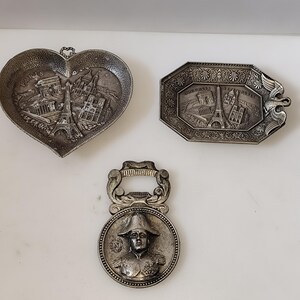 Könnte beinhalten: Ein Set aus drei silberfarbenen Metallgegenständen: eine herzförmige Schale mit geprägten Pariser Wahrzeichen, ein achteckiges Tablett mit Adler-Motiv und Pariser Monumenten sowie ein runder Flaschenöffner mit einer Büste von Napoleon Bonaparte.