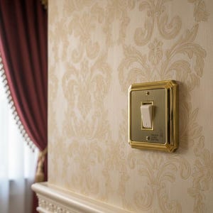以下が含まれることがあります： クリーム色のダマスク柄の壁紙が貼られた壁に、白いトグルが付いた金色のライトスイッチが取り付けられています。背景には、金のトリムが付いた赤いカーテンと白いモールディングが見えます。