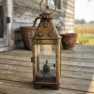 Peut inclure: Une lanterne en métal patiné jaune et brun, dotée de parois en verre transparent, d'une poignée circulaire sur le dessus et d'un mécanisme de brûleur à huile central. Le sommet pyramidal présente des orifices de ventilation circulaires.