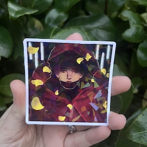 May include: Square sticker featuring a person in a red hooded cloak, surrounded by yellow petal shapes. The artwork has a digital painting style with a dark color palette and a white border. The sticker is held in a hand against a background of green leaves.