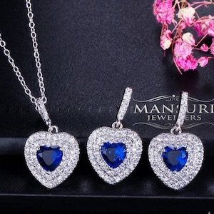 Puede incluir: Un conjunto de joyería en tono plateado que consta de un collar y dos pendientes colgantes. Cada pieza presenta una piedra azul central en forma de corazón rodeada por un doble halo de pequeñas piedras claras. El collar incluye una cadena fina.