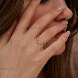 May include: A rose gold V-shaped ring adorned with small, clear gemstones. The ring is worn on a person's finger, with the hand partially covering the face. The ring's design is a delicate, modern style, suitable for everyday wear or special occasions.
