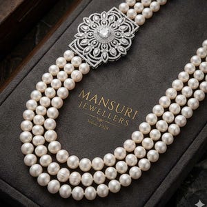 Op de afbeelding: Een parelketting met drie strengen en een decoratieve, met diamanten bezette sluiting. De sluiting heeft een centrale diamant omgeven door een ingewikkeld bloemmotief. De ketting wordt gepresenteerd in een grijze sieradendoos. De tekst "MANSURI JEWELLERS Since 1928" is zichtbaar.