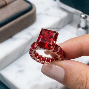 May include: A large, red gemstone ring with a rectangular cut. The ring features a band of smaller red stones and a gold setting. The ring is held up against a white marble surface.