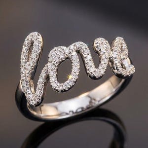Peut inclure: Une bague de couleur argentée présentant le mot love en écriture cursive sur le dessus. Les lettres sont incrustées de nombreuses petites pierres claires et scintillantes qui captent la lumière, sur un anneau en métal poli, lisse et brillant.