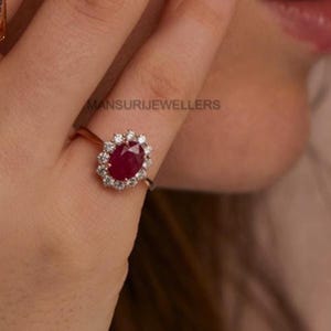 May include: A ring featuring a central oval-cut deep red ruby surrounded by a halo of small round white diamonds set on a thin metallic band. The text MANSURIJEWELLERS is visible in the background behind the ring setting.