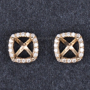 Puede incluir: Dos engastes de pendientes cuadrados dorados, cada uno con un halo de pequeñas piedras preciosas transparentes. El centro de cada engaste presenta un diseño en forma de X. Los pendientes se muestran sobre una superficie gris oscuro.