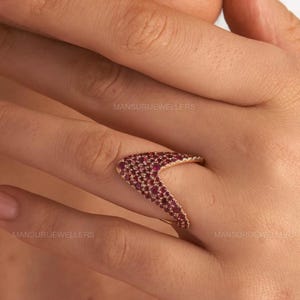 Könnte beinhalten: Ein goldfarbener Ring mit V-förmigem Design, besetzt mit zahlreichen kleinen, tiefroten Edelsteinen. Der Ring ist vor einem hautfarbenen Hintergrund platziert, der die Farbe und Form des Schmucks hervorhebt. Der Text "MANSURIJEWELLERS" ist sichtbar.