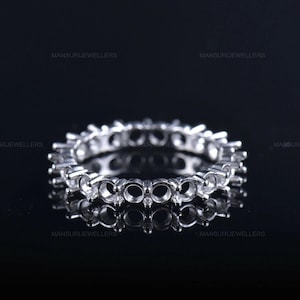 Könnte beinhalten: Ein silberner Eternity-Ring mit mehreren runden Fassungen für Edelsteine. Der Ring ist hochreflektierend und hat eine polierte Oberfläche. Der Ring wird auf einer schwarzen Oberfläche präsentiert. Der Text "MANSURIJEWELLERS" ist sichtbar.