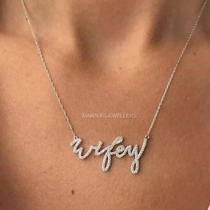 以下が含まれることがあります： 筆記体で「wifey」と書かれたシルバーのネックレス。ネックレスは小さな輝く石で飾られています。「MANSURIJEWELLERS」というブランド名がネックレスの上に印刷されています。