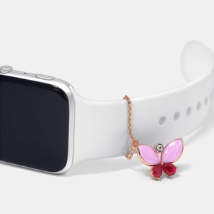May include: A rose gold butterfly charm with pink and red details, attached to a white smartwatch strap. The butterfly features a small diamond. The watch has a silver case.