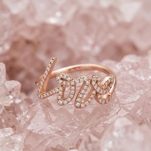 May include: Rose gold ring spelling out the word "love" in cursive script. The letters are encrusted with small, sparkling diamonds. The ring is set against a soft pink, crystalline background, creating a romantic and elegant aesthetic.