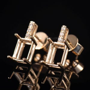 May include: Close-up of two gold-colored stud earrings. Each earring features a rectangular setting with four prongs and a vertical bar with small, clear gemstones. The earrings are set against a dark, reflective background.