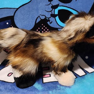 May include: A plush toy fox with a fluffy tail and fur in shades of brown, cream, and black. The toy has black ears and paws, and is lying on a blue patterned surface.