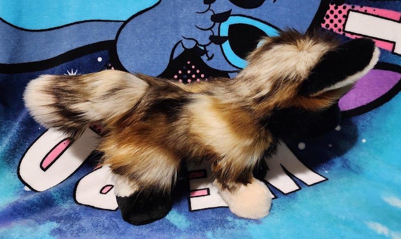 May include: A plush toy fox with a fluffy tail and fur in shades of brown, cream, and black. The toy has black ears and paws, and is lying on a blue patterned surface.
