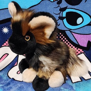 May include: A plush African wild dog toy with a black face, ears, and paws, and a multi-colored body of brown, tan, and white. The toy has a golden-colored eye and is sitting on a blue background.