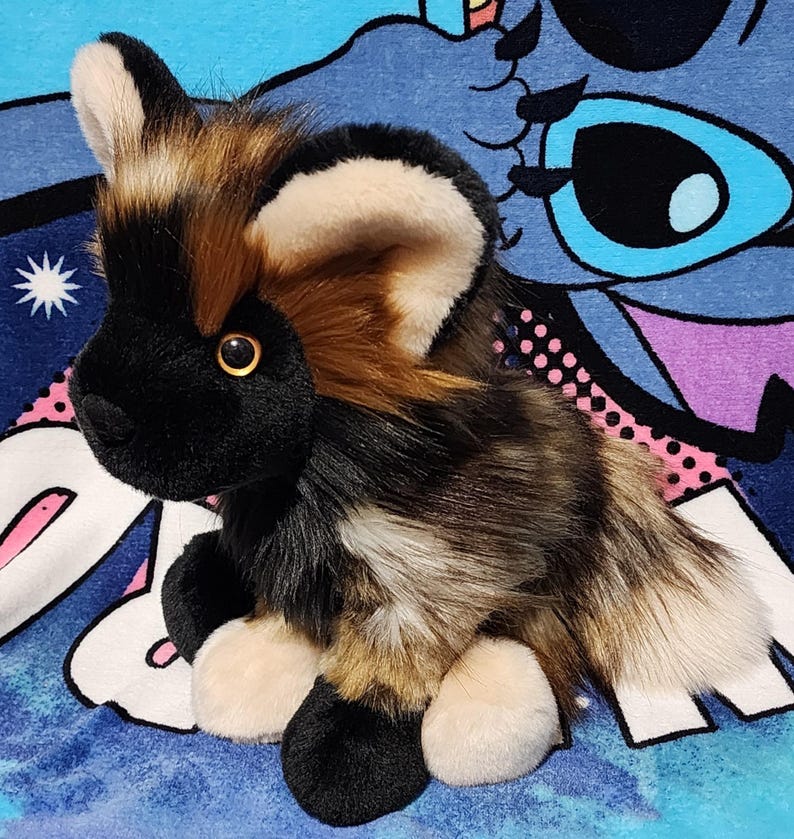 May include: A plush African wild dog toy with a black face, ears, and paws, and a multi-colored body of brown, tan, and white. The toy has a golden-colored eye and is sitting on a blue background.