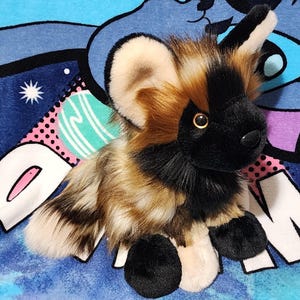 May include: A plush African wild dog toy with a black face, ears, and paws, and a tan and brown body. The toy has large, round ears and a soft, furry texture. It is sitting on a blue patterned blanket.