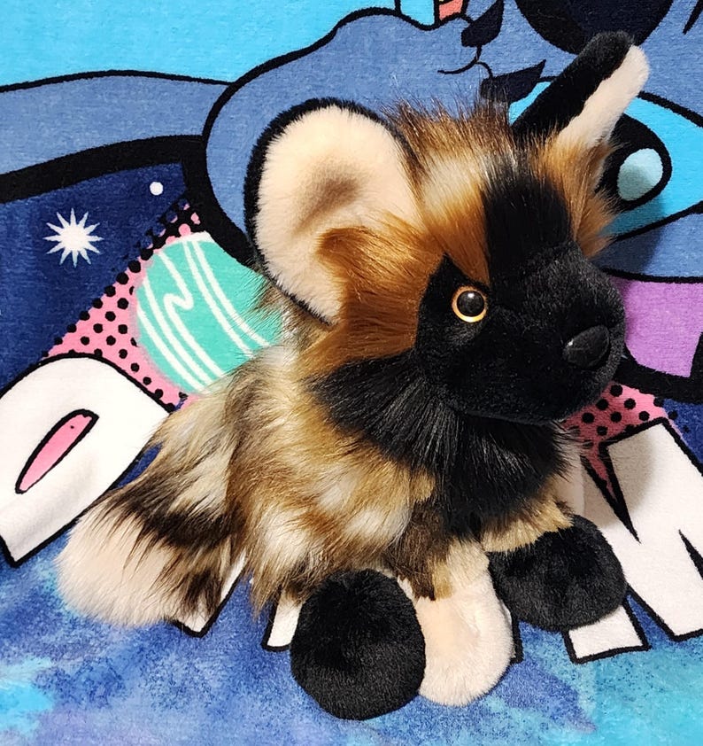 May include: A plush African wild dog toy with a black face, ears, and paws, and a tan and brown body. The toy has large, round ears and a soft, furry texture. It is sitting on a blue patterned blanket.