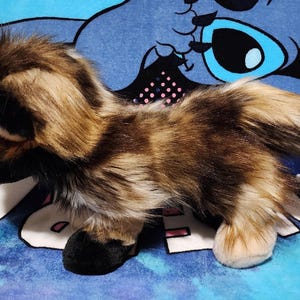 May include: A plush toy wolf with brown, tan, and black fur. The toy has black ears and paws, and a fluffy tail with a white tip. The toy is on a blue and purple patterned surface.