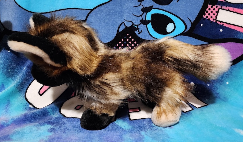 May include: A plush toy wolf with brown, tan, and black fur. The toy has black ears and paws, and a fluffy tail with a white tip. The toy is on a blue and purple patterned surface.