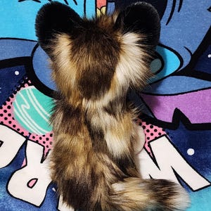 May include: A plush toy in the shape of a fox, featuring a mix of brown, tan, and black faux fur. The fox has large, black-tipped ears and a fluffy tail. The toy is set against a colorful, patterned background.